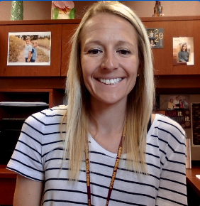 A smiling modern career counselor with straight blonde hair sits at a desk in a bright office, holding a clipboard labeled “Career Counseling.” Shelves with books, plants, and office supplies are visible behind her, creating a welcoming professional setting.